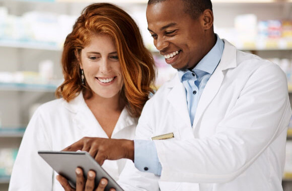 Two smiling healthcare professionals, a man and a woman, wearing white lab coats and looking at a tablet in a laboratory setting
